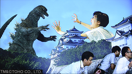 名古屋城の向こう側でゴジラと巨大な女性が戦い、名古屋城の手前で男性3名が逃げている写真
