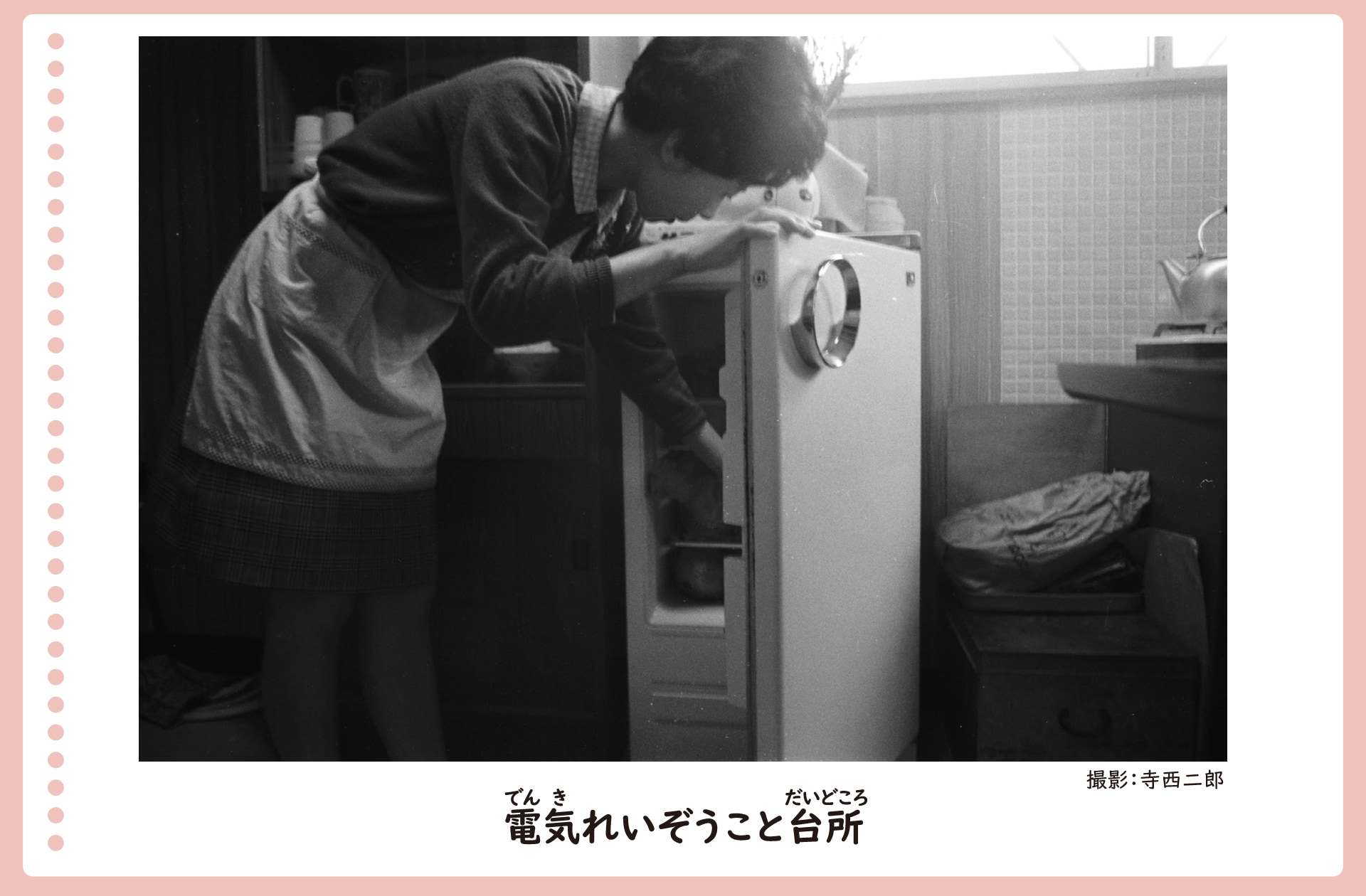 電気冷蔵庫がおかれた台所の風景。昭和40年。エプロンをつけた女性が冷蔵庫をひらき、かがんでなかの物をとっている。