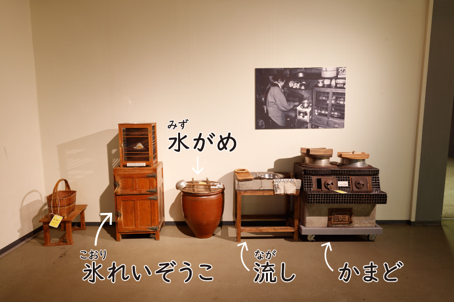 電化まえの台所を再現した様子。木でできた氷冷蔵庫と、水をためる水がめと、石でできた食器を洗う流しと、木を燃やしてごはんをたくかまどがならんでいる。